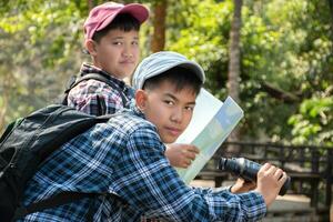 Asian boy in plaid shirt and cap doing the summer activity at local national park by watching birds, fish, insects, animals, trees, flower by using map and binoculars, soft and selective focus. photo