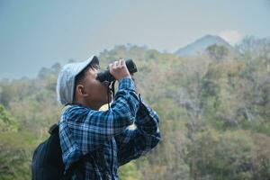 Asian boy in plaid shirt and cap doing the summer activity at local national park by watching birds, fish, insects, animals, trees, flower by using map and binoculars, soft and selective focus. photo
