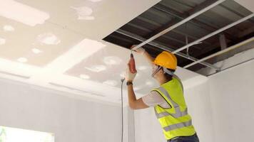 asiático construcción trabajador en la seguridad ropa y trabajo guantes es fijación el paneles de yeso techo a el metal marco utilizando un eléctrico destornillador en el techo cubrir foto