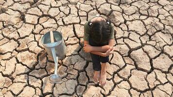 Sad little girl sitting with a watering can on dry ground. Water crisis, Concept hope and drought. photo