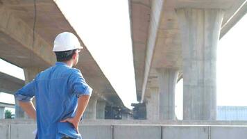 asiático joven profesional ingeniero trabajador en protector casco y planos papel en mano inspeccionando construcción proyecto un la carretera Autopista ese es hecho a construcción sitio. foto