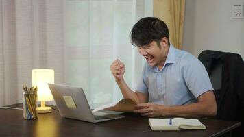 Asian man receives happy news in the mail. In the home, he sits at a table with a laptop, holds an envelope in his hands, reads, smiles. photo