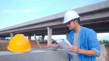 asiático civil ingeniero utilizando ordenador portátil y Plano es inspeccionando construcción proyecto un la carretera Autopista ese es hecho a construcción sitio. foto