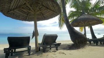 View of empty chaise-longue near native sun umbrella and palm trees against blue water, Mauritius Island video