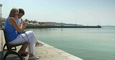 Man and woman using pad sitting on waterfront bench video