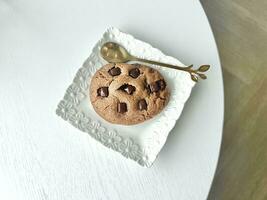 Chocolate chip cookie on a white plate with a golden spoon on white wooden table, top view photo