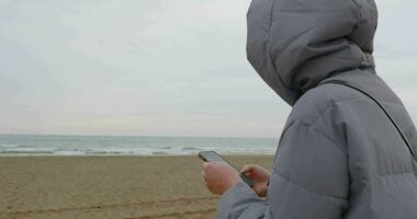 Woman in jacket surfing net on mobile near the sea on dull cold day video