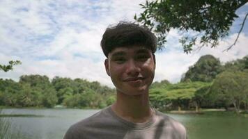 portrait asiatique Masculin adolescent dans décontractée robe souriant en dessous de le ombre de le arbre dans le la nature parc. video