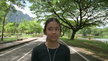 Happy adorable teen girl walking on the street and listening music with earphone in nature public park during summer. video