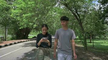 asiático adorable niña utilizando móvil teléfono inteligente y caminando juntos con novio en el naturaleza parque. video