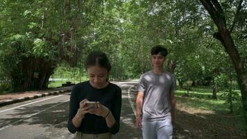 asiatique adolescent frère dans tenue de sport marcher Suivant le sien sœur qui en utilisant mobile téléphone dans Publique parc pendant été. video