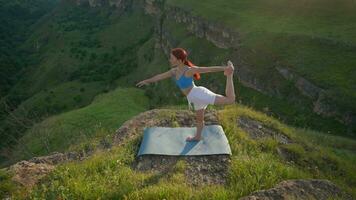 atlético mujer haciendo yoga poses en parte superior de un montaña durante puesta de sol. Deportes niña trenes y lo hace extensión ejercicios en el montañas. sano estilo de vida, zenismo, rutina de ejercicio concepto. lento movimiento. video