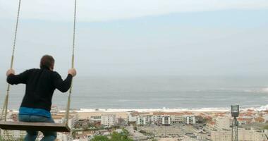 hombre balanceo en el colina con vista a recurso pueblo y océano. nazaré en Portugal video