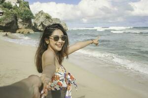 un contento joven asiático mujer es sonriente a el cámara y señalando a el playa en gunungkidul, Indonesia. foto