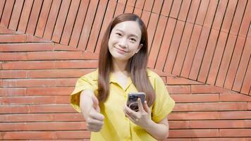 ung asiatisk kvinna innehav en smartphone och höjning henne hand i ett excellent gest ser på de kamera video