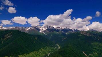 Panoramic aerial view of summer mountain valley landscape with green forest, snowy peaks, mountain river and sky with clouds video