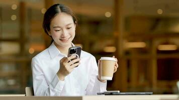 Smiling Asian woman looking at mobile phone talking about work while holding coffee in the morning video