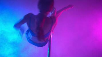 Athletic woman pole dancer performs a stunt number on a pole in a smoky room in neon light. video