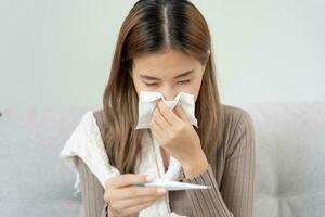 Young Asian woman having high fever while checking body temperature, female sneezing and runny nose with seasonal influenza, allergic, digital thermometer, virus, coronavirus, illness, respiratory photo