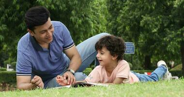 Video of father talking to his cute son while lying on grass in garden drawing pictures with him.