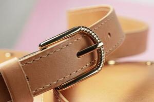 Part of an expensive brown leather item with stitching and a metal buckle on a pink background. photo