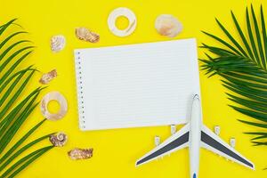 Palm branches with frame of shells and notepad, plane on summer yellow background. Vacation, travel concept, flat lay top view, minimal exotic concept. Copy space photo