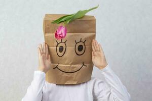 The boy has a cardboard box above his head with a funny smiley face drawn on it. The tulip is on top. photo