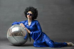 espectacular mujer en un brillante acampanado traje y afro peluca poses con un disco pelota. disco estilo desde el setenta o años ochenta foto