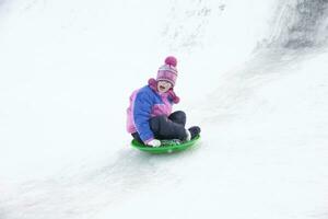un niña en un invierno chaqueta gritando se mueve abajo desde un nieve diapositiva foto