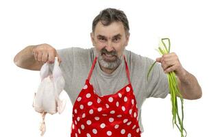 An elderly bearded male cook in a red polka-dot apron holds a raw chicken and a bunch of onions on a white background. Cook with chicken. photo