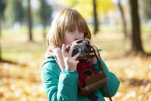 pequeño Pelirrojo niña con un retro cámara en el otoño parque. niño fotógrafo. foto