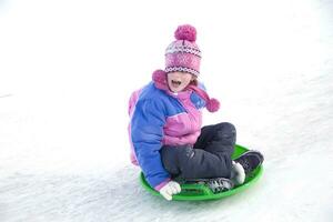 un niña en un invierno chaqueta gritando se mueve abajo desde un nieve diapositiva foto