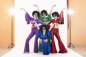 A group of girls in colorful flared suits and afro wigs pose in a photo studio. Disco style from the eighties or seventies.