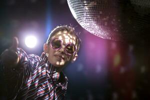 A young boy in stylish glasses shows a class sign against the background of light rays from a disco ball. Children's disco, party. photo