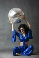 A woman in a seventies flared suit with a disco ball in an afro wig on a gray background. photo