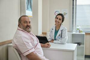 hermosa hembra médico en uniforme y blanco masculino paciente sonriente y mirando a cámara, salud chequeo cita a un trabajando escritorio, sano dieta clínica hospital. foto