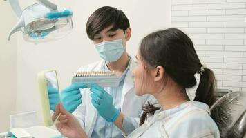 Aziatisch mannetje tandarts legt uit orthodontisch zorg en bleken naar een jong vrouw geduldig met implantaat kunstgebit kleur schaduw in tandheelkundig kliniek, tanden hygiëne, en professioneel gezondheidszorg dokter in een ziekenhuis. video