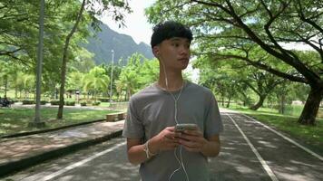 Asian handsome teen student listening to music while standing under the shade of big tree in city park. video