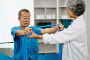 asiático fisioterapeuta Ayudar mayor hombre paciente extensión brazo durante ejercicio correcto con pesa en mano durante formación mano en cama en clínica foto