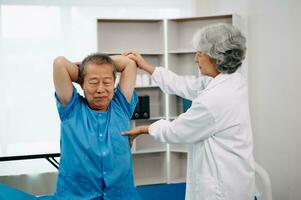 asiático fisioterapeuta Ayudar mayor hombre paciente extensión brazo durante ejercicio correcto con pesa en mano durante formación mano en cama en clínica foto