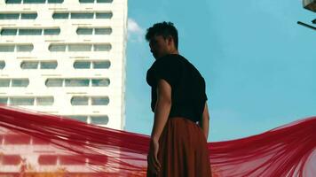 an Asian man standing in front of a building with a blue sky and red cloth flying video