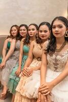 a group of Asian women sitting together on a white bed while wearing dresses and makeup during a party at a friend's house photo