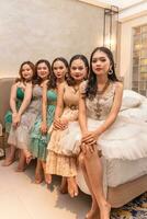 a group of Asian women sitting together on a white bed while wearing dresses and makeup during a party at a friend's house photo