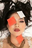 an Asian woman with a gold necklace falls asleep with a condom wrapper on her face in a hotel photo