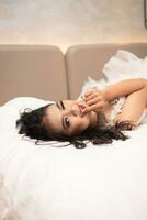 an Asian woman with black hair biting her fingers with a horny expression while sleeping on a white bed photo