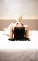 un asiático mujer en un blanco vestir es acostado en un blanco cama con su negro pelo suelto en un hotel foto