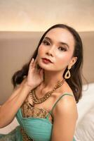 un asiático mujer con un oro collar y pendientes es posando hermosamente en un hotel habitación antes de cena foto