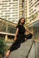 an Asian man poses on a sloping rock while wearing black around an abandoned building photo