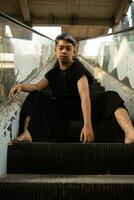 un asiático hombre en negro ropa es sentado en un roto y abandonado ascensor en un antiguo edificio foto