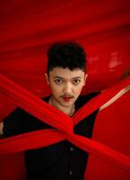 an Asian man stands in front of a red x-shaped cloth and holds it with both hands photo